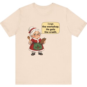 K&ouml;nnte beinhalten: Ein hellbeiges T-Shirt mit einer Cartoon-Illustration einer &auml;lteren Person in einem rot-wei&szlig;en Festtagsoutfit mit gr&uuml;ner Sch&uuml;rze, die einen Holzl&ouml;ffel und ein Klemmbrett h&auml;lt, mit einer Sprechblase, die besagt, I run the workshop. He gets the credit.