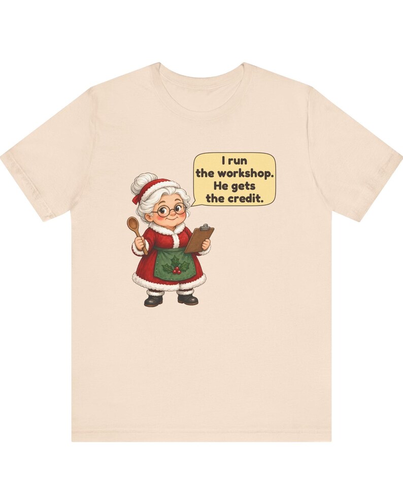 K&ouml;nnte beinhalten: Ein hellbeiges T-Shirt mit einer Cartoon-Illustration einer &auml;lteren Person in einem rot-wei&szlig;en Festtagsoutfit mit gr&uuml;ner Sch&uuml;rze, die einen Holzl&ouml;ffel und ein Klemmbrett h&auml;lt, mit einer Sprechblase, die besagt, I run the workshop. He gets the credit.