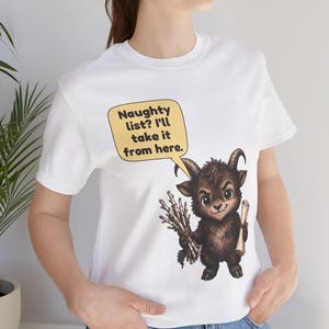 Puede incluir: Una camiseta blanca de manga corta con el gr&aacute;fico de una criatura marr&oacute;n con cuernos que sostiene un manojo de palos y un pergamino. Un bocadillo amarillo sobre la criatura contiene el texto: Naughty list? I'll take it from here.