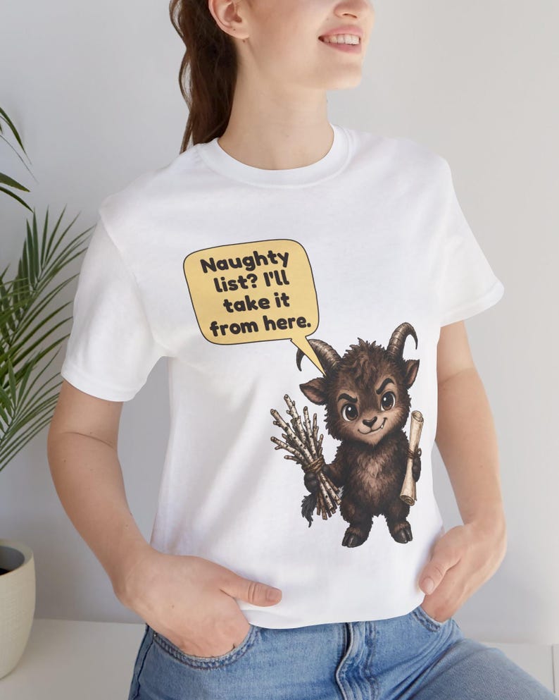 Puede incluir: Una camiseta blanca de manga corta con el gr&aacute;fico de una criatura marr&oacute;n con cuernos que sostiene un manojo de palos y un pergamino. Un bocadillo amarillo sobre la criatura contiene el texto: Naughty list? I'll take it from here.
