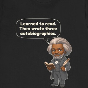 Puede incluir: Una camiseta negra con una ilustraci&oacute;n estilo caricatura de Frederick Douglass sosteniendo un libro y una pluma. Un globo de texto sobre &eacute;l contiene el texto: Learned to read. Then wrote three autobiographies. El dise&ntilde;o est&aacute; centrado.