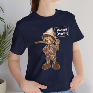 K&ouml;nnte beinhalten: Ein kurz&auml;rmeliges T-Shirt in Marineblau mit einer Grafik einer Holzpuppe mit langer Nase, die ein gemustertes Outfit und einen Hut tr&auml;gt. Eine Sprechblase neben der Figur enth&auml;lt den Text, Honest! (Mostly.)