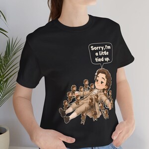 Op de afbeelding: Een zwart t-shirt met een afbeelding van een groot persoon in 18e-eeuwse kleding die op de grond ligt, omringd door vele kleine mensen die touwen en ladders gebruiken om hen vast te binden. Een tekstballon boven de figuur zegt, Sorry, I'm a little tied up.