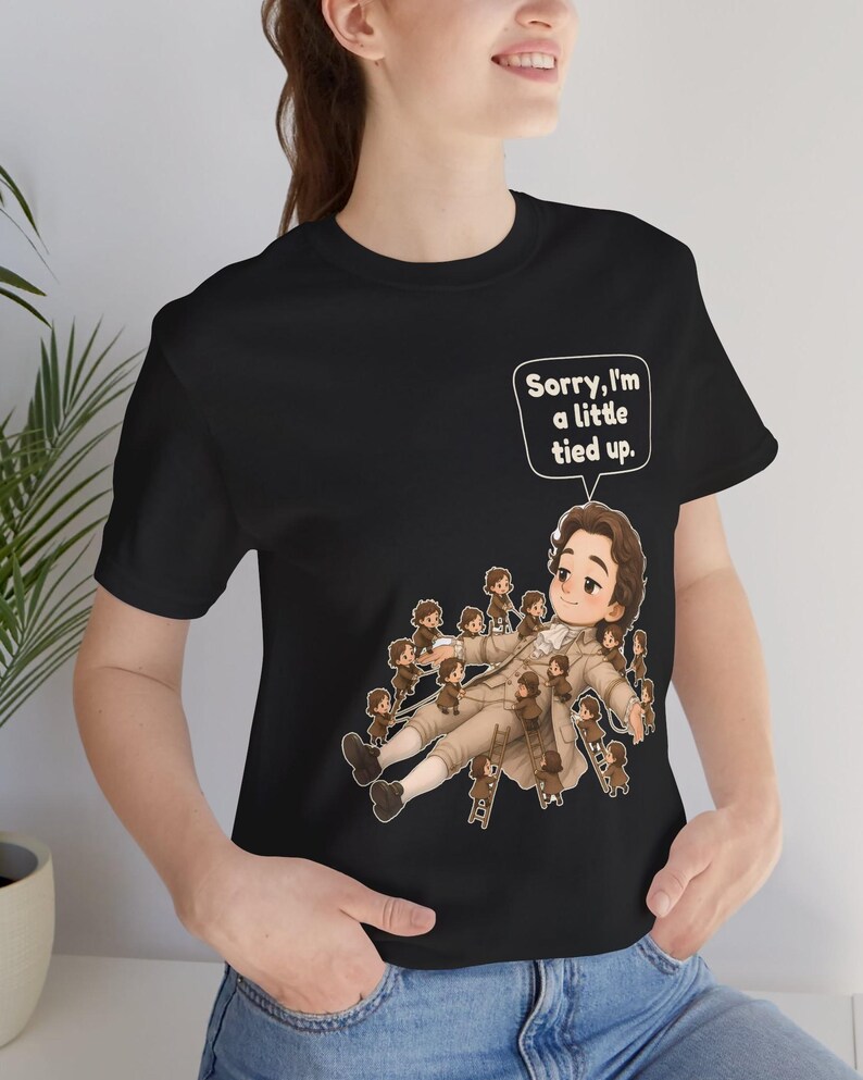 Op de afbeelding: Een zwart t-shirt met een afbeelding van een groot persoon in 18e-eeuwse kleding die op de grond ligt, omringd door vele kleine mensen die touwen en ladders gebruiken om hen vast te binden. Een tekstballon boven de figuur zegt, Sorry, I'm a little tied up.