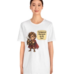 May include: A white t-shirt featuring a cartoon illustration of a warrior in bronze armor and a red cape, holding a plumed helmet. A speech bubble next to the character contains the text: Conquered the world by 30. You?