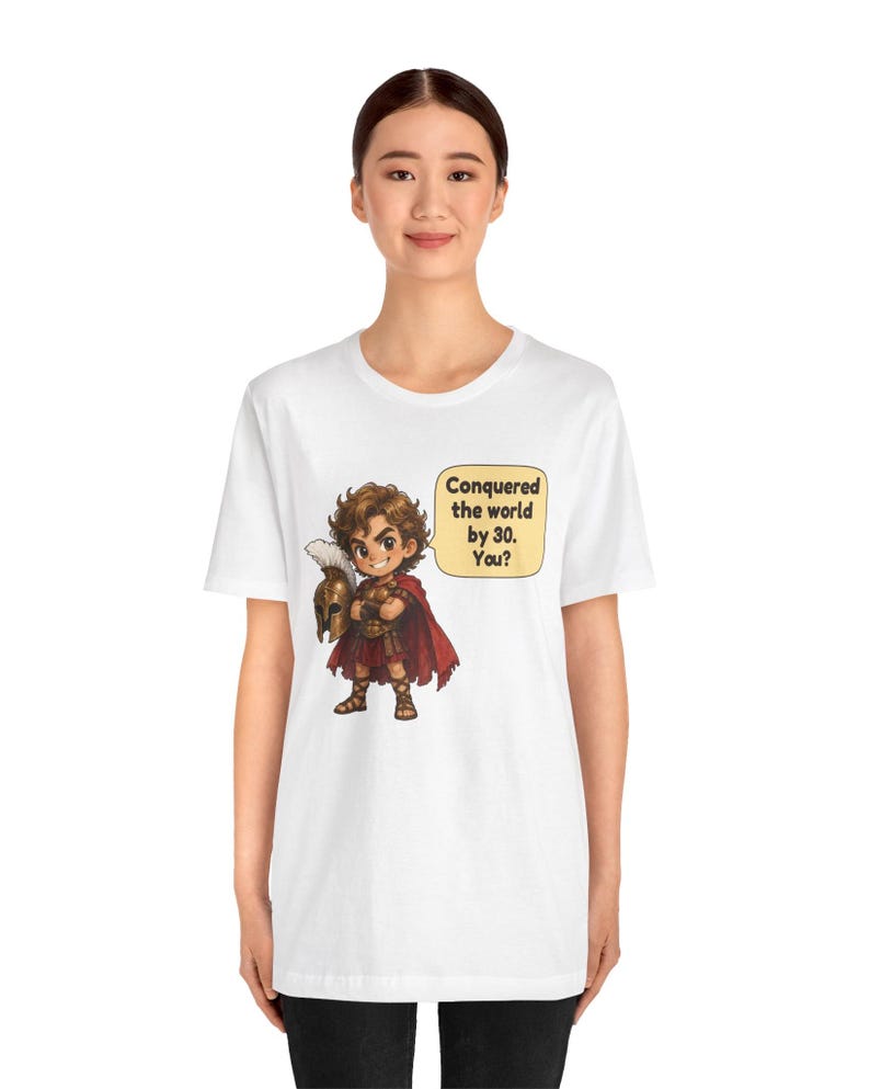 May include: A white t-shirt featuring a cartoon illustration of a warrior in bronze armor and a red cape, holding a plumed helmet. A speech bubble next to the character contains the text: Conquered the world by 30. You?