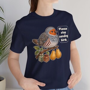 K&ouml;nnte beinhalten: Ein marineblaues T-Shirt mit einer Grafik eines runden Vogels, der auf einem Ast mit zwei Birnen und zwei kleinen Geschenkkartons sitzt. Der Vogel hat eine Sprechblase mit dem Text: "Please stop sending birds." Das Motiv ist mittig platziert.