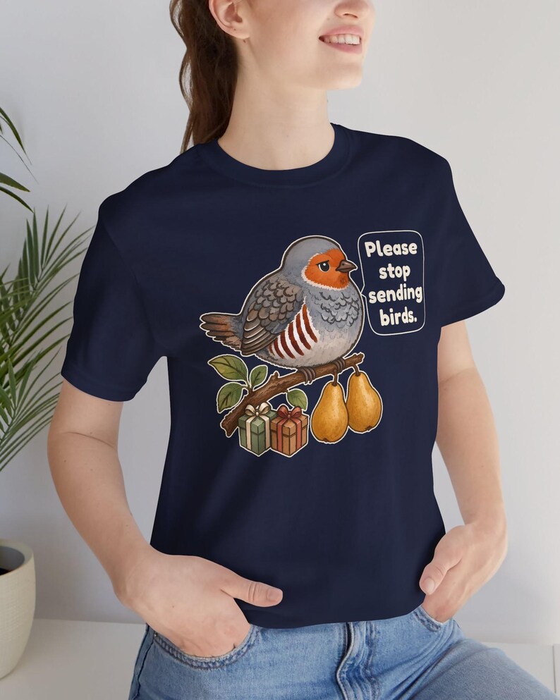 K&ouml;nnte beinhalten: Ein marineblaues T-Shirt mit einer Grafik eines runden Vogels, der auf einem Ast mit zwei Birnen und zwei kleinen Geschenkkartons sitzt. Der Vogel hat eine Sprechblase mit dem Text: "Please stop sending birds." Das Motiv ist mittig platziert.