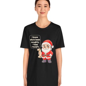 May include: A black t-shirt featuring a cartoon Santa Claus holding a scroll. A speech bubble next to Santa contains the text: I know who's been naughty. I keep receipts. Santa wears a red suit with white trim, a red hat, and dark boots.