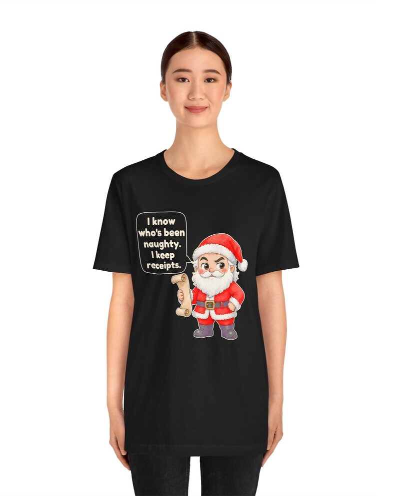 May include: A black t-shirt featuring a cartoon Santa Claus holding a scroll. A speech bubble next to Santa contains the text: I know who's been naughty. I keep receipts. Santa wears a red suit with white trim, a red hat, and dark boots.
