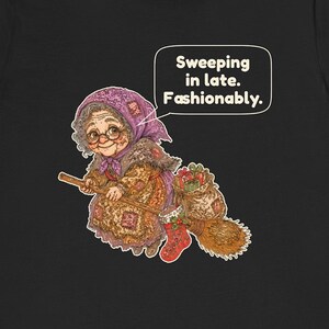 Puede incluir: Una camiseta negra con el gr&aacute;fico de una persona mayor con pelo gris, gafas y pa&ntilde;uelo morado montada en una escoba. La persona viste un vestido estampado y lleva un saco de regalos y un calcet&iacute;n rojo. El texto dice: Sweeping in late. Fashionably.