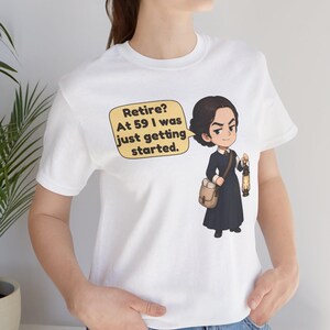 May include: A white t-shirt featuring a cartoon illustration of a person in a dark dress carrying a satchel and a lantern. A speech bubble next to the figure contains the text: Retire? At 59 I was just getting started.