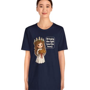 May include: A navy blue t-shirt featuring a graphic of a character wearing a white robe and a crown of candles with greenery, holding a tray of buns. A speech bubble next to the character contains the text: Bringing the light (and the buns).