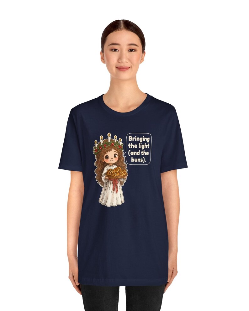 May include: A navy blue t-shirt featuring a graphic of a character wearing a white robe and a crown of candles with greenery, holding a tray of buns. A speech bubble next to the character contains the text: Bringing the light (and the buns).