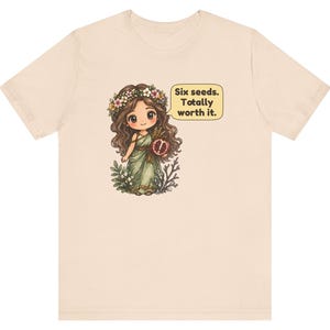 May include: A light beige t-shirt featuring a graphic of a cartoon character with long brown hair, a flower crown, and a green dress. The character holds a pomegranate and has a speech bubble that reads: Six seeds. Totally worth it.