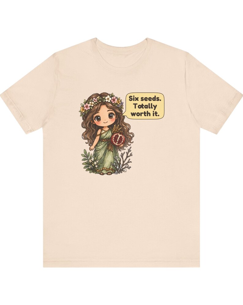 May include: A light beige t-shirt featuring a graphic of a cartoon character with long brown hair, a flower crown, and a green dress. The character holds a pomegranate and has a speech bubble that reads: Six seeds. Totally worth it.