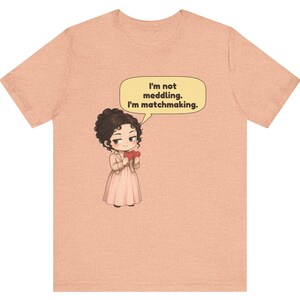 K&ouml;nnte beinhalten: Ein pfirsichfarbenes Kurzarm-T-Shirt mit einer Cartoon-Illustration einer Person mit dunklem, lockigem Haar, die ein helles Kleid tr&auml;gt und zwei rote Herzen h&auml;lt. Eine Sprechblase dar&uuml;ber enth&auml;lt den Text: I'm not meddling. I'm matchmaking.