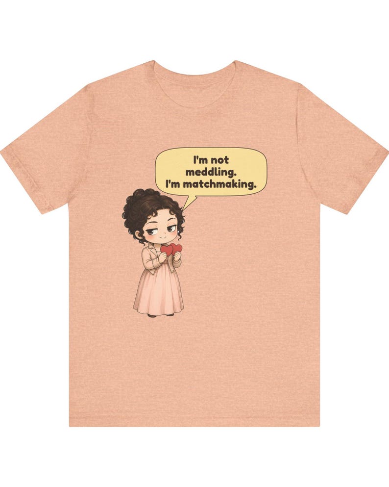 K&ouml;nnte beinhalten: Ein pfirsichfarbenes Kurzarm-T-Shirt mit einer Cartoon-Illustration einer Person mit dunklem, lockigem Haar, die ein helles Kleid tr&auml;gt und zwei rote Herzen h&auml;lt. Eine Sprechblase dar&uuml;ber enth&auml;lt den Text: I'm not meddling. I'm matchmaking.