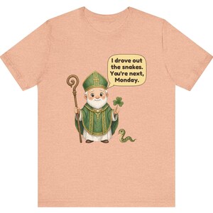 Puede incluir: Una camiseta color melocot&oacute;n con una ilustraci&oacute;n de dibujos animados de una figura con vestiduras religiosas verdes que sostiene un bast&oacute;n de madera y un tr&eacute;bol, con una peque&ntilde;a serpiente y un globo de texto: I drove out the snakes. You're next, Monday.