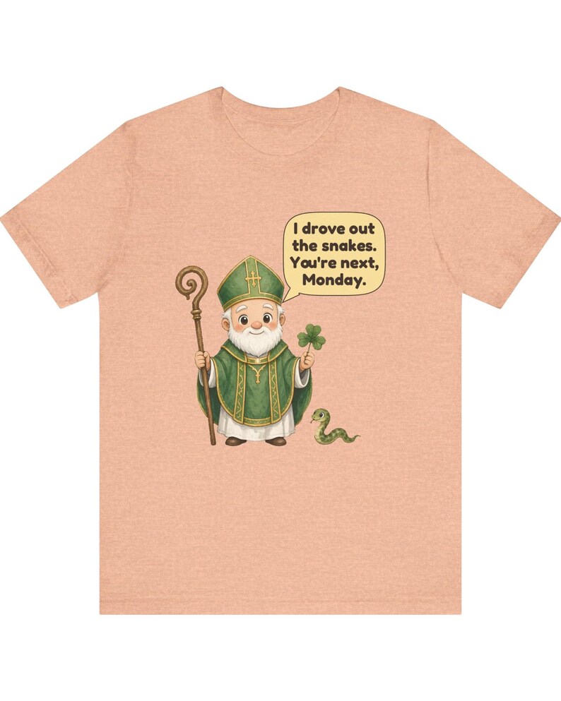 Puede incluir: Una camiseta color melocot&oacute;n con una ilustraci&oacute;n de dibujos animados de una figura con vestiduras religiosas verdes que sostiene un bast&oacute;n de madera y un tr&eacute;bol, con una peque&ntilde;a serpiente y un globo de texto: I drove out the snakes. You're next, Monday.