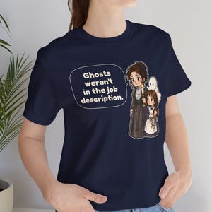 Op de afbeelding: Een marineblauw t-shirt met een afbeelding van een vrouw en een kind die een kaars vasthouden, vergezeld door een klein spookje. Een tekstballon ernaast bevat de tekst: Ghosts weren't in the job description. Het t-shirt heeft korte mouwen.