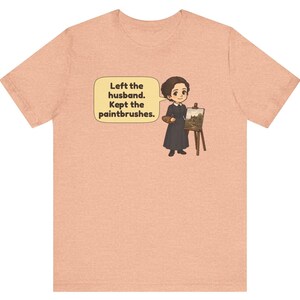 May include: A peach-colored short-sleeve t-shirt featuring a cartoon illustration of a person in a dark dress standing by an easel with a painting. A speech bubble contains the text: Left the husband. Kept the paintbrushes.