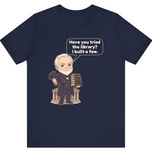 May include: A navy blue t-shirt featuring a cartoon illustration of a white-haired man in a dark suit holding a stack of books. A speech bubble above him contains the text, "Have you tried the library? I built a few." The man stands before a building.