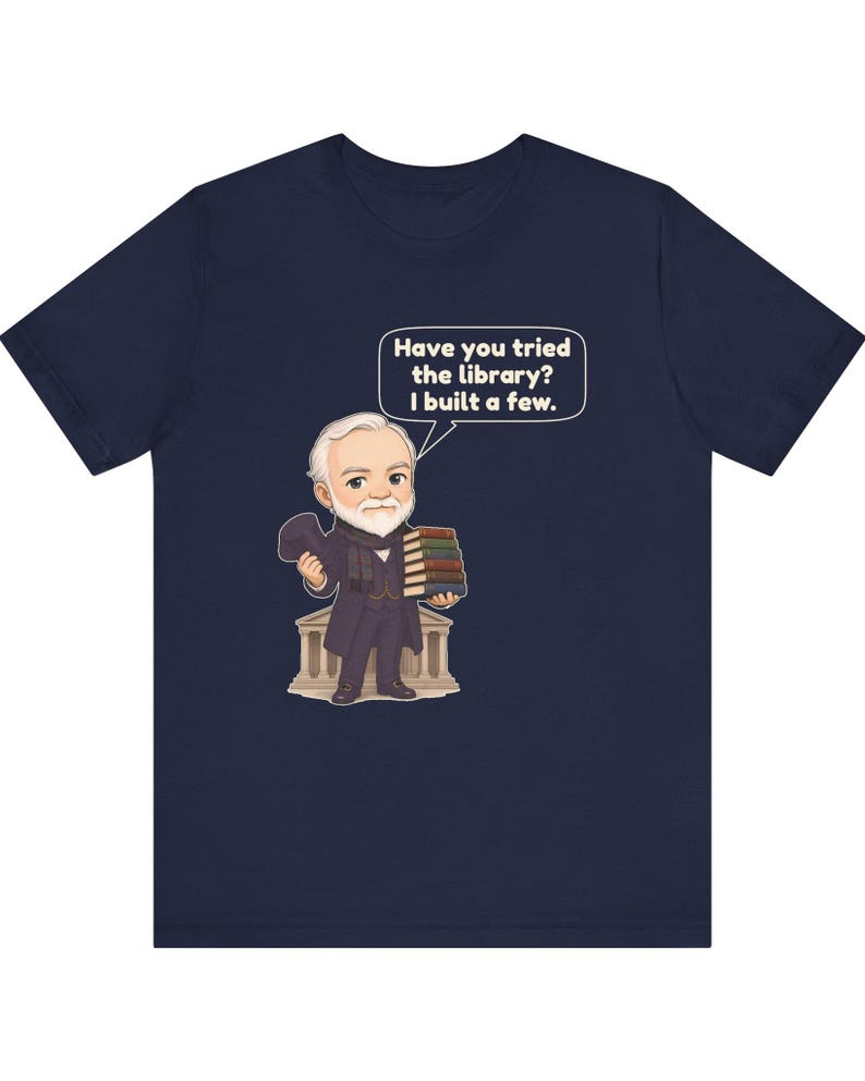 May include: A navy blue t-shirt featuring a cartoon illustration of a white-haired man in a dark suit holding a stack of books. A speech bubble above him contains the text, "Have you tried the library? I built a few." The man stands before a building.