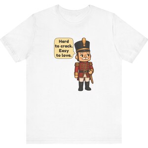Pu&ograve; includere: Una t-shirt bianca con un soldatino schiaccianoci in stile cartoon, che indossa un'uniforme rossa con dettagli dorati, stivali neri e un alto cappello nero. Un fumetto accanto alla figura contiene il testo: Hard to crack. Easy to love.