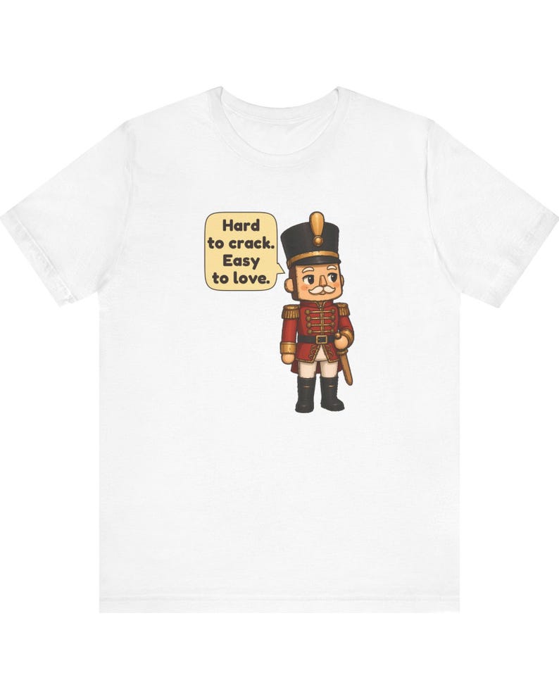 Pu&ograve; includere: Una t-shirt bianca con un soldatino schiaccianoci in stile cartoon, che indossa un'uniforme rossa con dettagli dorati, stivali neri e un alto cappello nero. Un fumetto accanto alla figura contiene il testo: Hard to crack. Easy to love.