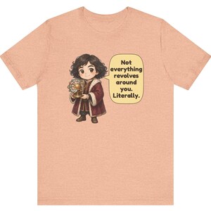 May include: A peach-colored t-shirt featuring a cartoon illustration of a person in historical clothing holding a model of the solar system. A speech bubble next to the figure contains the text: Not everything revolves around you. Literally.