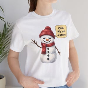 Pode incluir: Uma t-shirt branca de manga curta com o gr&aacute;fico de um boneco de neve a usar um gorro e um cachecol de malha vermelha. O boneco de neve tem um nariz de cenoura, bra&ccedil;os de pau e um bal&atilde;o com o texto: Chill. It's just a phase.