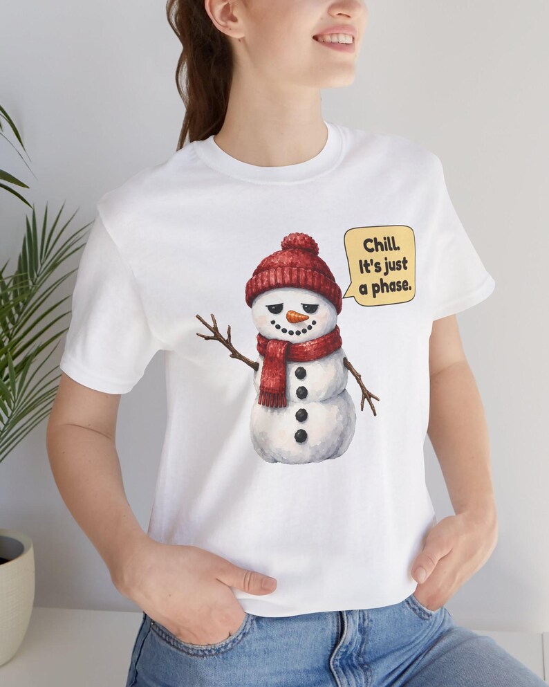 Pode incluir: Uma t-shirt branca de manga curta com o gr&aacute;fico de um boneco de neve a usar um gorro e um cachecol de malha vermelha. O boneco de neve tem um nariz de cenoura, bra&ccedil;os de pau e um bal&atilde;o com o texto: Chill. It's just a phase.