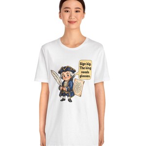 May include: A white t-shirt featuring a cartoon illustration of a historical figure in a blue coat and tricorn hat holding a large white quill. A speech bubble next to the character contains the text: Sign big. The king needs glasses.