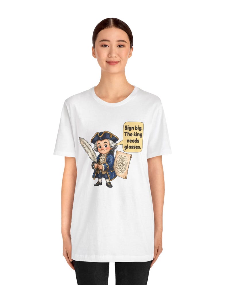 May include: A white t-shirt featuring a cartoon illustration of a historical figure in a blue coat and tricorn hat holding a large white quill. A speech bubble next to the character contains the text: Sign big. The king needs glasses.