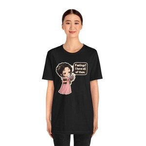 K&ouml;nnte beinhalten: Ein schwarzes T-Shirt mit einer Grafik einer Zeichentrickfigur mit dunklem Haar in einem rosa Kleid, die ein Buch und Blumen h&auml;lt. Eine Sprechblase neben der Figur enth&auml;lt den Text: Feelings? I have all of them.