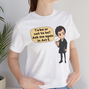以下が含まれることがあります： 白い半袖Tシャツで、暗い服を着たキャラクターが頭蓋骨を持っているグラフィックが描かれています。黄色い吹き出しには「To be or not to be? Ask me again in Act 5.」というテキストが含まれています。デザインは衣服の前面中央に配置されています。
