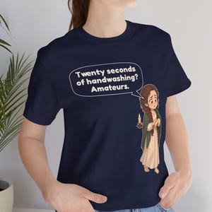 Op de afbeelding: Een marineblauw t-shirt met een cartoonillustratie van een persoon met een lange vlecht die een kaars vasthoudt. Een tekstballon boven het personage bevat de tekst: Twenty seconds of handwashing? Amateurs.