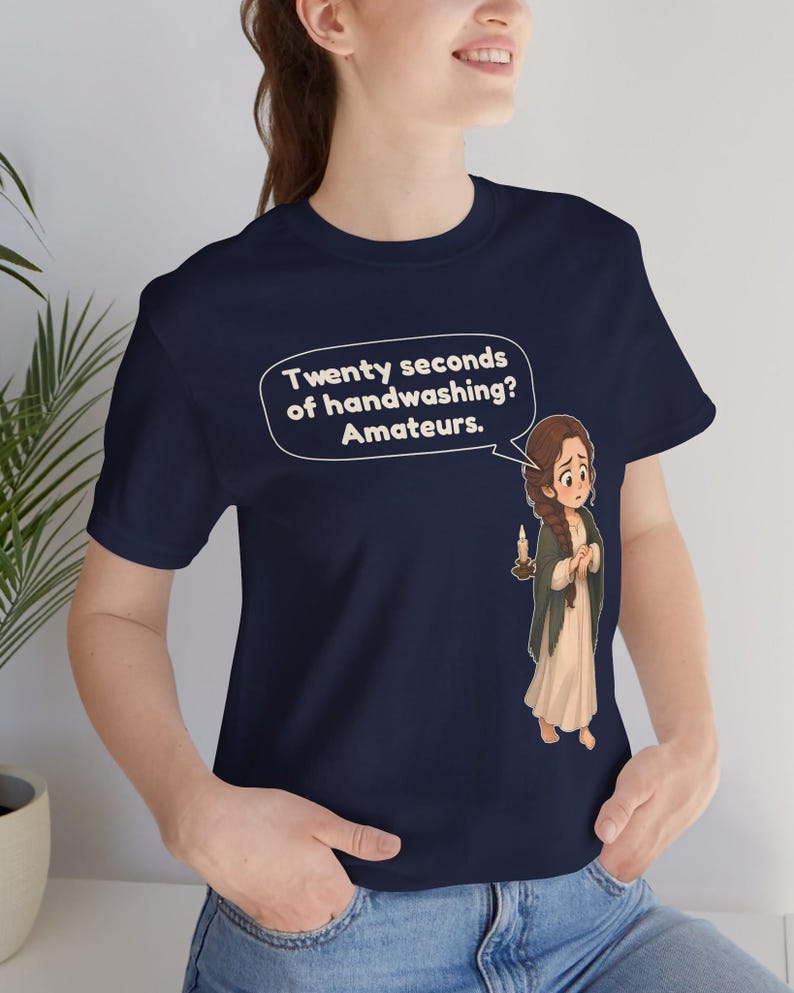 Op de afbeelding: Een marineblauw t-shirt met een cartoonillustratie van een persoon met een lange vlecht die een kaars vasthoudt. Een tekstballon boven het personage bevat de tekst: Twenty seconds of handwashing? Amateurs.