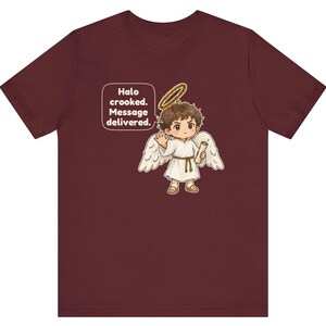 May include: A maroon short-sleeve t-shirt featuring a graphic of a cartoon angel with brown hair, white wings, and a gold halo. The angel holds a scroll and waves. A speech bubble contains the text: Halo crooked. Message delivered.