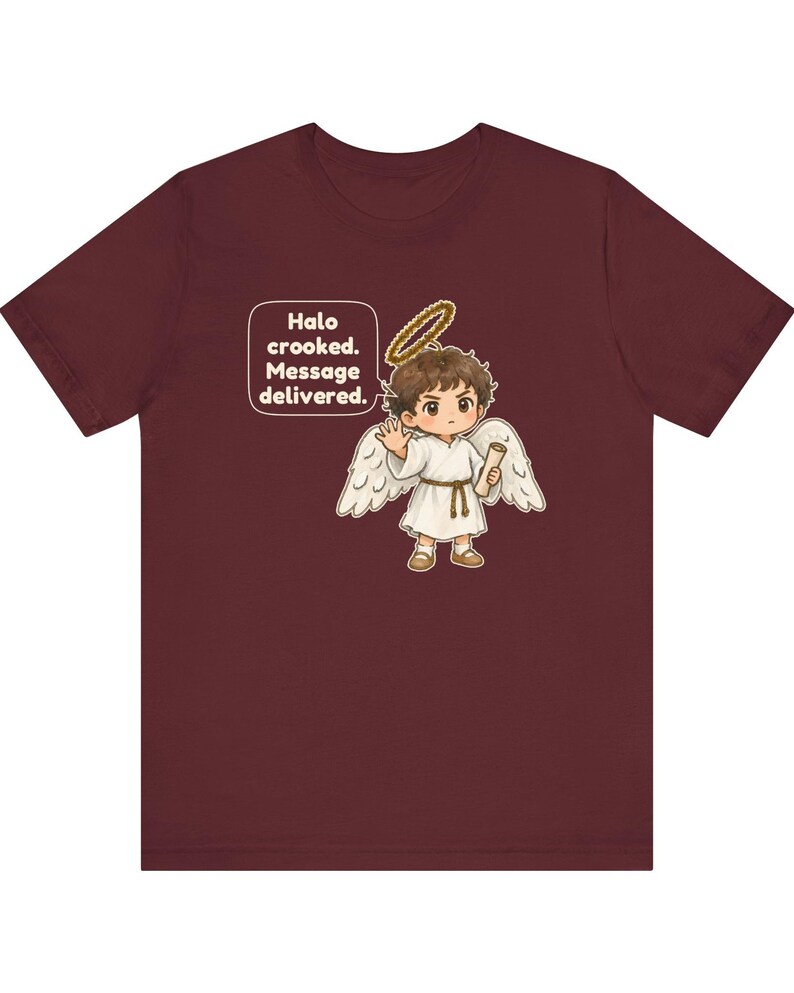 May include: A maroon short-sleeve t-shirt featuring a graphic of a cartoon angel with brown hair, white wings, and a gold halo. The angel holds a scroll and waves. A speech bubble contains the text: Halo crooked. Message delivered.