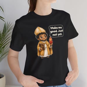 May include: A black t-shirt featuring a cartoon illustration of a bishop in gold vestments holding a book and a flaming heart. A speech bubble above the figure contains the text: Make me good. Just not yet.