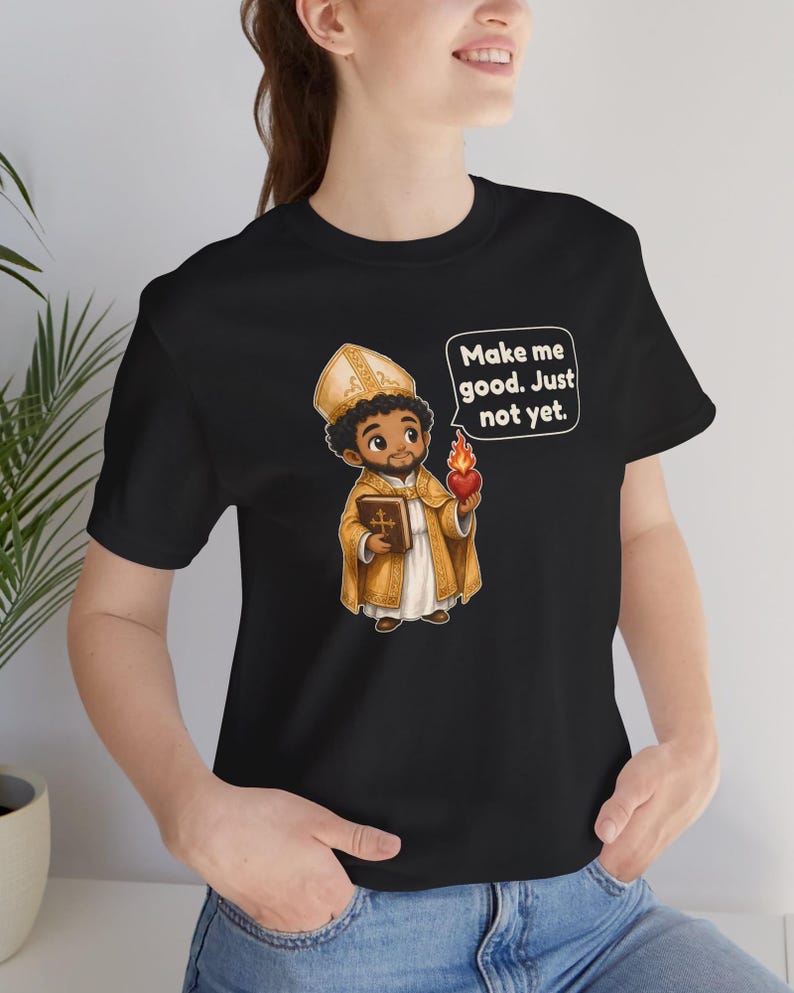 May include: A black t-shirt featuring a cartoon illustration of a bishop in gold vestments holding a book and a flaming heart. A speech bubble above the figure contains the text: Make me good. Just not yet.