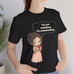 K&ouml;nnte beinhalten: Ein schwarzes T-Shirt mit einer Cartoon-Illustration einer Person mit braunen lockigen Haaren, die eine beige Jacke und ein rosa Kleid tr&auml;gt und zwei rote Herzen h&auml;lt. Eine Sprechblase enth&auml;lt den Text: I'm not meddling. I'm matchmaking.