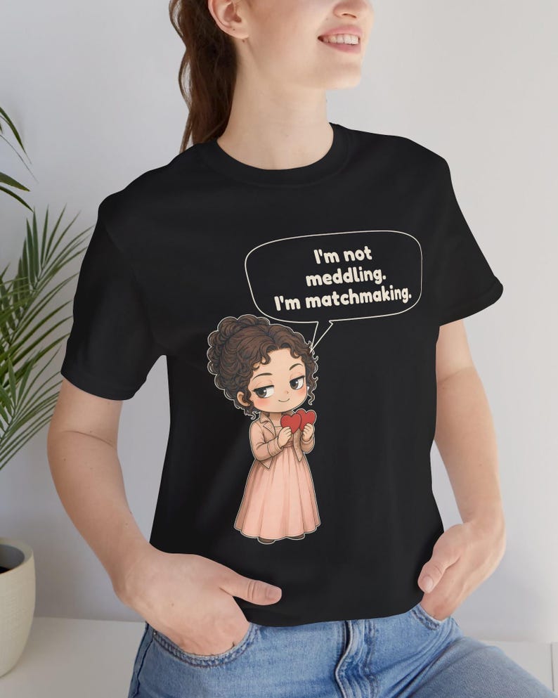 K&ouml;nnte beinhalten: Ein schwarzes T-Shirt mit einer Cartoon-Illustration einer Person mit braunen lockigen Haaren, die eine beige Jacke und ein rosa Kleid tr&auml;gt und zwei rote Herzen h&auml;lt. Eine Sprechblase enth&auml;lt den Text: I'm not meddling. I'm matchmaking.
