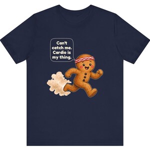 May include: A navy blue t-shirt featuring a graphic of a running gingerbread person wearing a red and white headband. A speech bubble above the character contains the text: Can't catch me. Cardio is my thing. A cloud of dust follows behind.