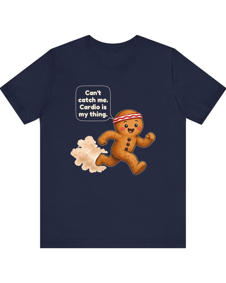 May include: A navy blue t-shirt featuring a graphic of a running gingerbread person wearing a red and white headband. A speech bubble above the character contains the text: Can't catch me. Cardio is my thing. A cloud of dust follows behind.