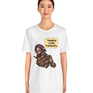 Puede incluir: Una camiseta blanca de manga corta con un gr&aacute;fico de una persona mayor con pa&ntilde;uelo y gafas, montada en una escoba. El gr&aacute;fico incluye un bocadillo con el texto: "Sweeping in late. Fashionably."