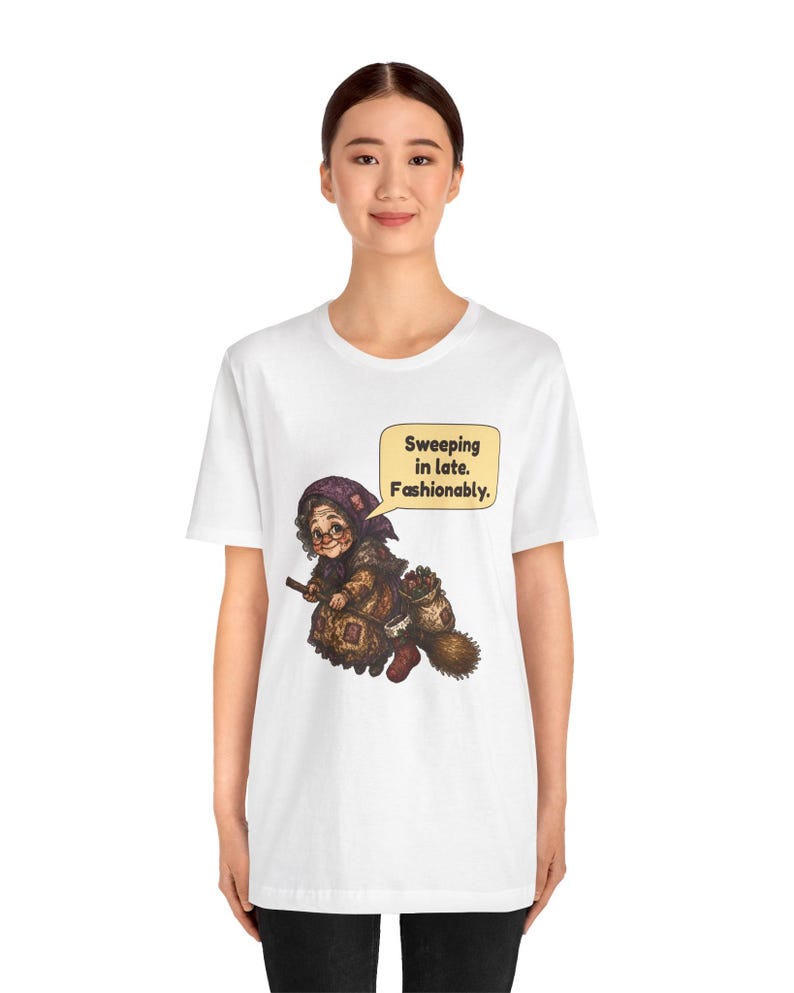 Puede incluir: Una camiseta blanca de manga corta con un gr&aacute;fico de una persona mayor con pa&ntilde;uelo y gafas, montada en una escoba. El gr&aacute;fico incluye un bocadillo con el texto: "Sweeping in late. Fashionably."