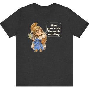 Puede incluir: Una camiseta gris oscuro con una ilustraci&oacute;n de dibujos animados de una figura con casco dorado y t&uacute;nica azul que sostiene una rama de olivo y un libro, con un b&uacute;ho en el hombro y un globo de texto: Show your work. The owl is watching.